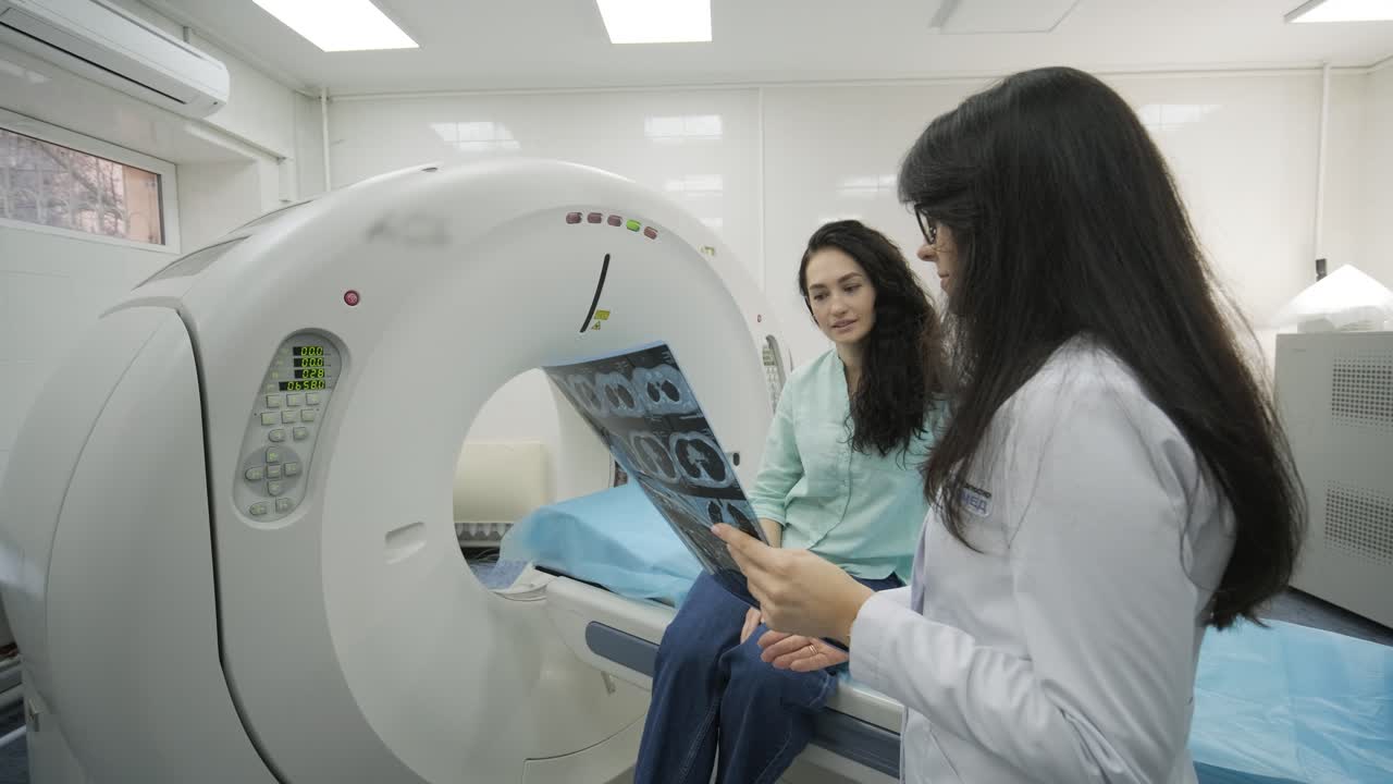 Woman doctor radiologist explains the results of CT scanning for young female patient, showing the snapshot with images, observing and analyzing CT scan in modern clinic beside modern CT scanner