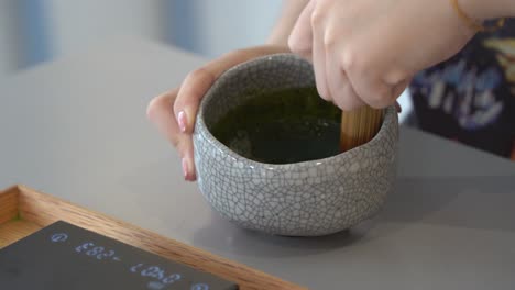 젊은 여성의 손이 손으로 만든 도자기 그릇에 말차 녹차를 휘젓고 대나무 휘젓기, 일본 전통 의식 말차, 전문 음료, 클로즈업 샷을 섞습니다.