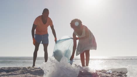 african american couple segregating waste with gloves on sunny beach