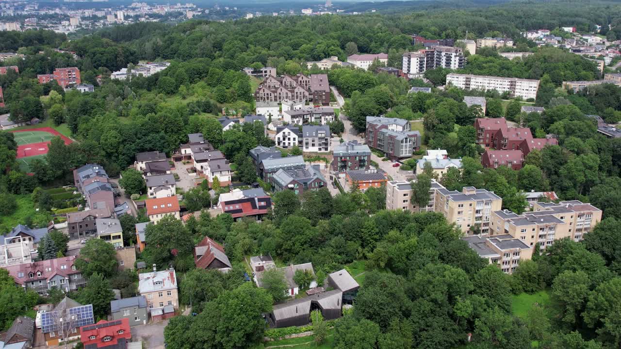 Drone aerial over the leafy Markučiai district in Vilnius, Lithuania, showing scattered homes and low-rise blocks surrounded by dense forest under a soft overcast sky