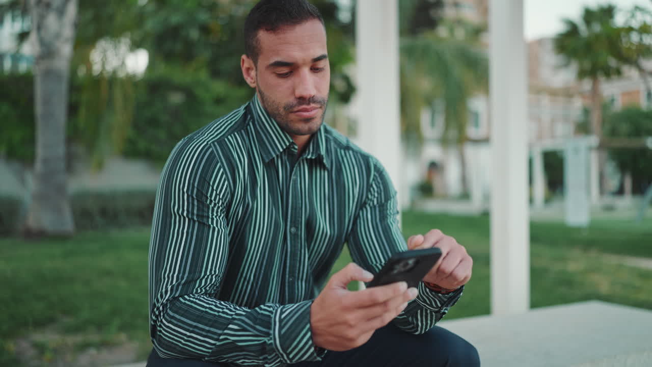 giovane che controlla lo smartphone seduto su una panchina all'aperto.