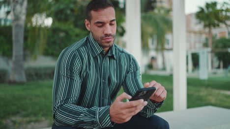 joven comprobando su teléfono inteligente sentado en un banco al aire libre.