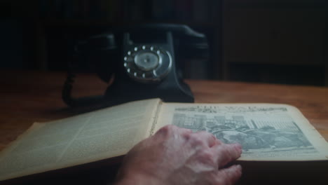 throwback history scene, retro glasses on an open encyclopedia, hand turns pages and answer call in antique telephone on the table. close up