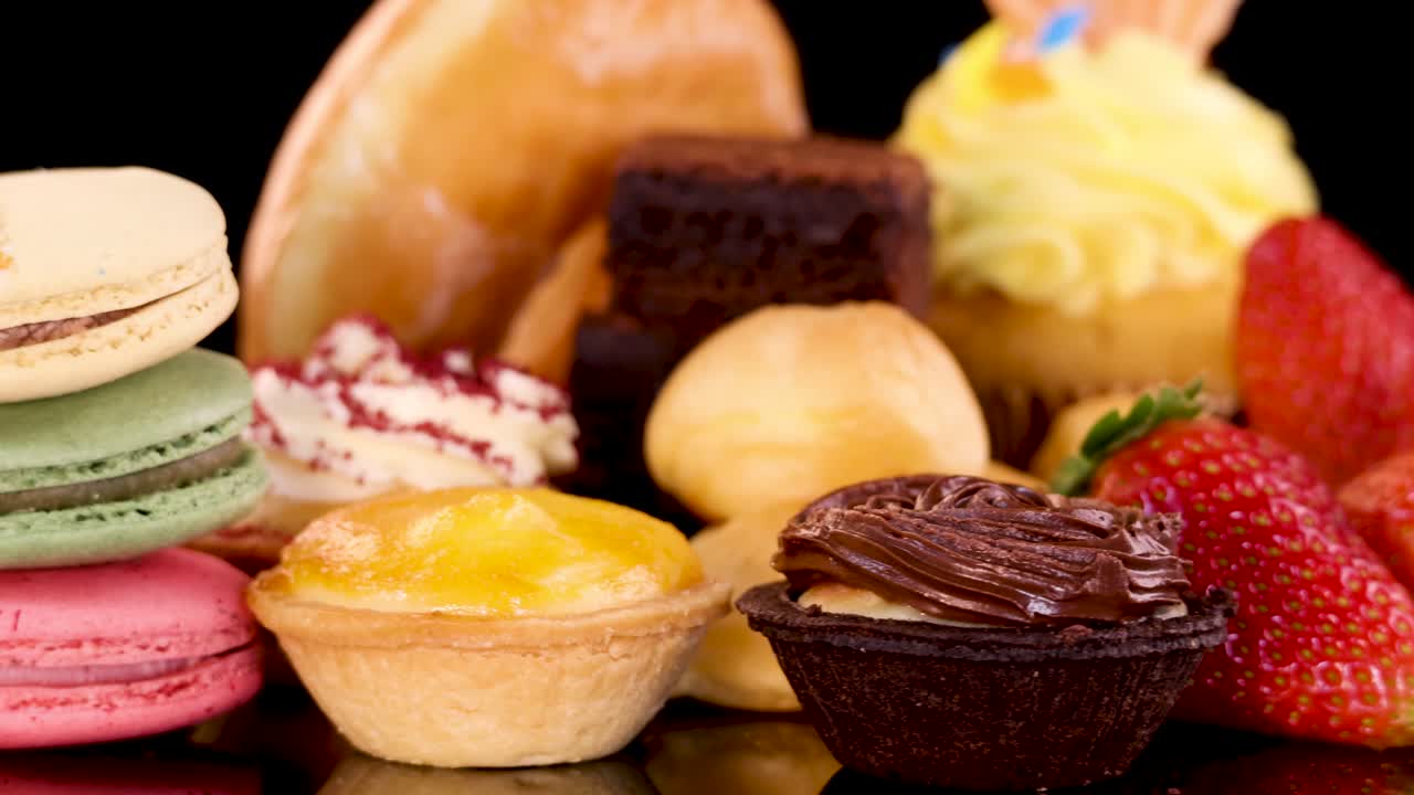 A rotating display of assorted bakery desserts including cupcakes, donuts, macarons, brownies, tarts, and strawberries, brightly lit against a black background with smooth camera movement