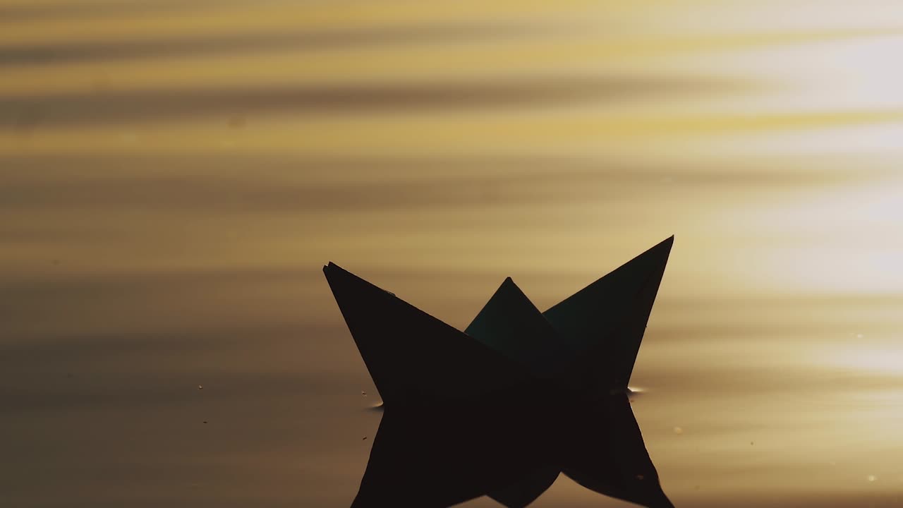 paper boat by handmade is floating on the waves of the river at sunset. The image of a paper ship is reflecting in the water in the evening