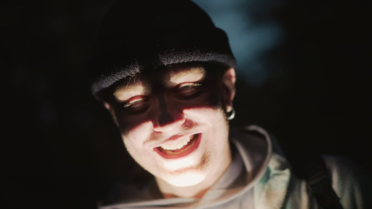 Volto caucasico illuminato da una lanterna, sorriso malizioso, ritratti che raffigurano un artista di strada, un comico da villaggio turistico, un esploratore urbano, un influencer notturno e un avventuriero amichevole, l'illuminazione frontale dal basso crea un effetto drammatico