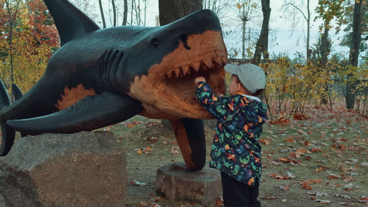 Lovely Caucasian boy standing near the wooden sculpture of a shark. Kid takes an acorn and shows it to the camera.