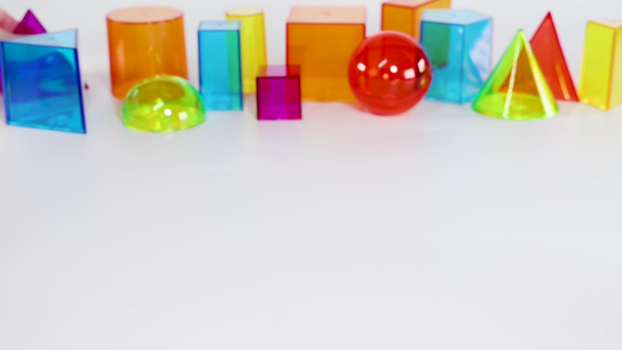 Hands interact with vibrant 3D geometric shapes on a white surface, highlighting prisms, spheres, and pyramids in bright lighting