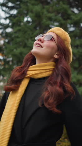 hermosa dama con cabello rizado rojo con boina amarilla, gafas de sol y silenciador se da la vuelta felizmente en un jardín vibrante rodeado de exuberante vegetación y follaje de otoño