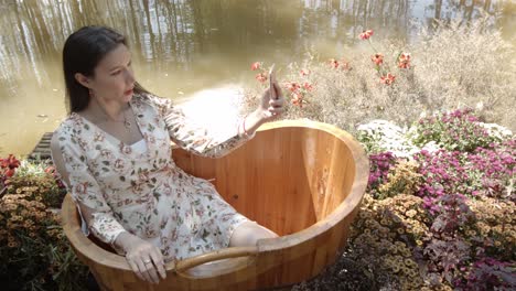 An-attractive-Asian-woman-sits-in-a-garden-tub-by-a-pond-and-takes-a-selfie-with-her-smartphone