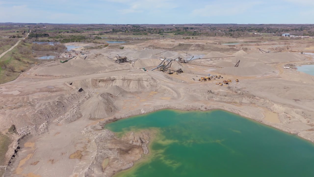 Drone video captures a sprawling gravel pit industry in Canada, featuring conveyor belts, trucks, turquoise pit lakes and rugged terrain under crisp spring daylight, with no people visible.