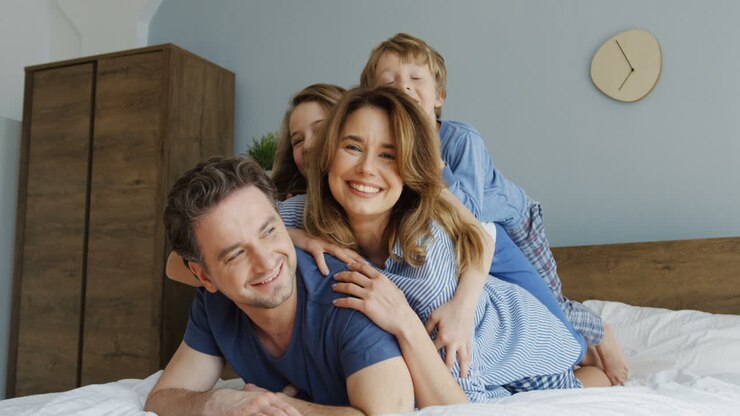 Cheerful parents and sons lying on the back of each other on the bed and smiling at the camera
