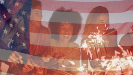 american flag waving against group of friends enjoying with sparklers at the beach