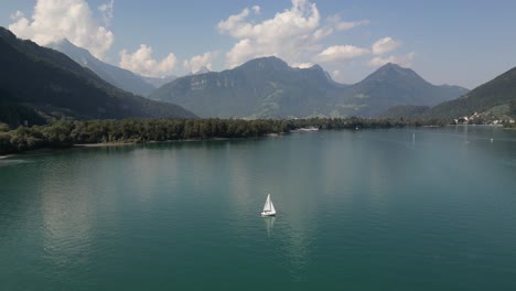 flying away from a small tiny sailing boat ship in the middle of massive lake pure water green great mountain heights in the background uneven sharp shapes clouds covering them natural selection place