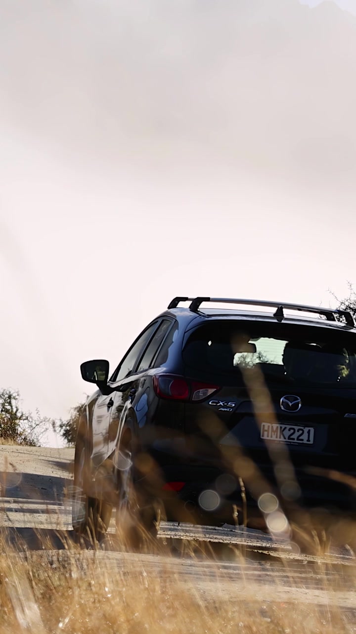Un SUV nero viaggia lungo una strada rurale tortuosa delimitata da erba secca e alberi sparsi, ripreso alla luce naturale del giorno con una ripresa in movimento costante da dietro.