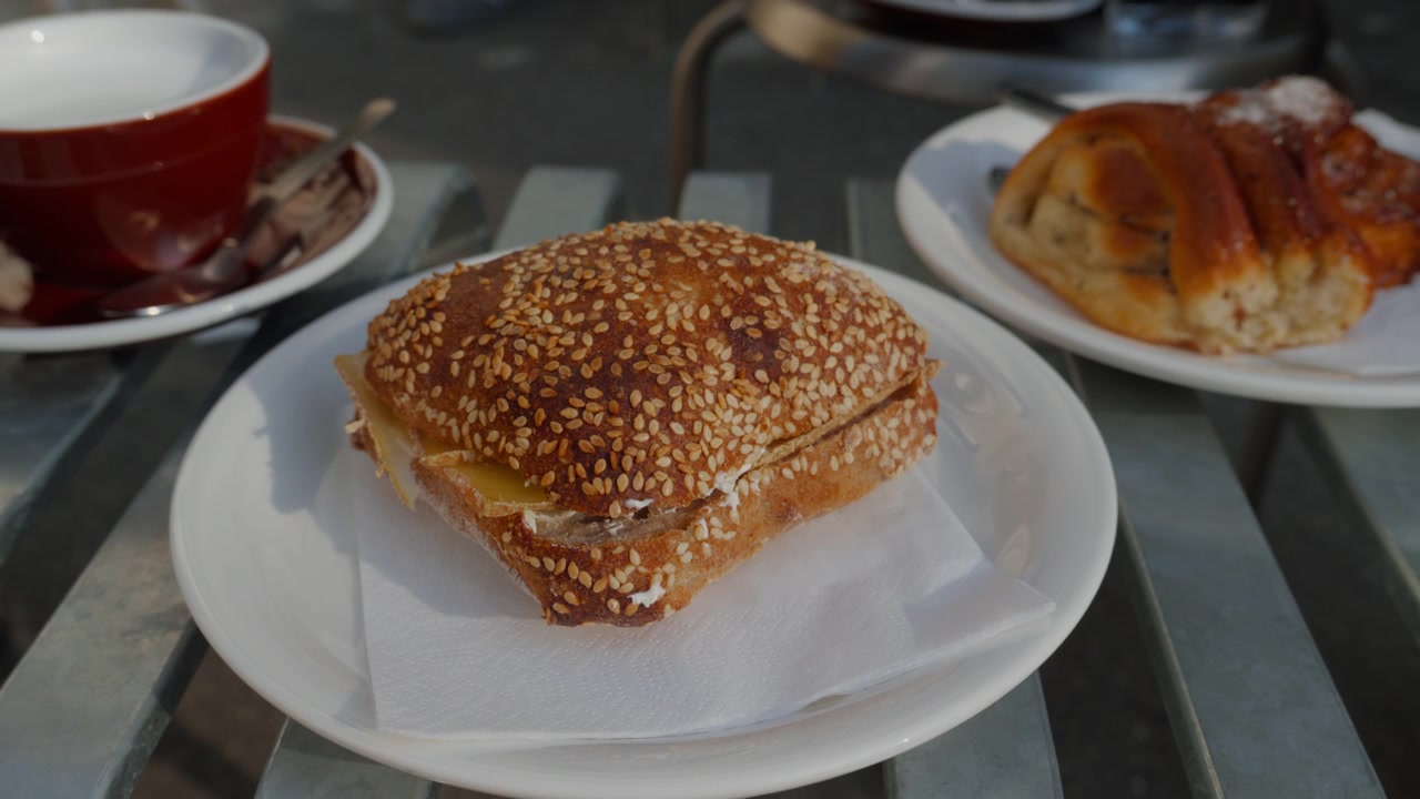 Close view of a fresh sesame seed roll filled with cheese on a white plate at a sunny cafe table, an appetising city breakfast.