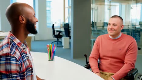 two men are talking at a desk. one man is sitting in a wheelchair.