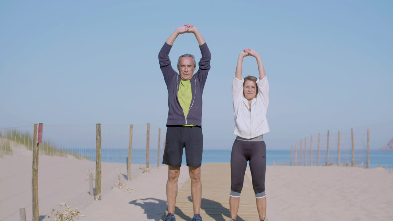 Focused elderly couple exercising on sandy beach on summer day