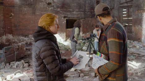 dos compañeros de producción hablando y leyendo un documento sobre la película en un edificio en ruinas mientras otros compañeros se preparan para la grabación