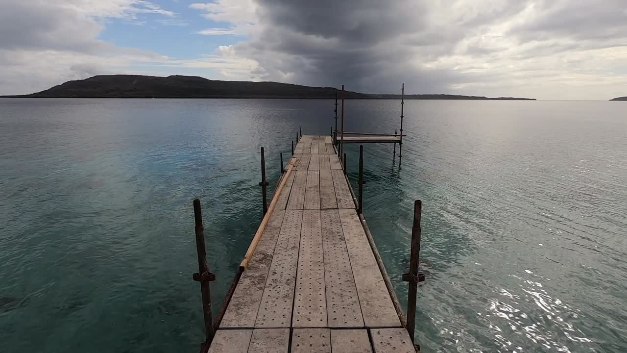 caminar sobre un muelle de madera en la costa del mar bajo un cielo dramático pov con una isla en el horizonte