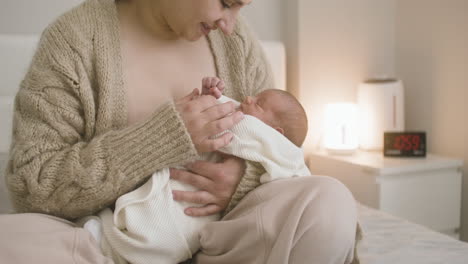 Front-View-Of-Brunette-Smiling-Woman-Sitting-On-The-Bed-Holding-Her-Baby-And-Caressing-Him-1