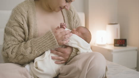 front view of brunette smiling woman sitting on the bed holding her baby and caressing him 1