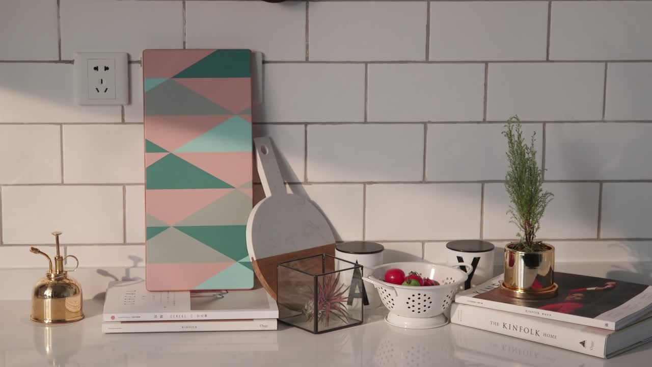 A sunlit kitchen counter with geometric cutting boards, potted herbs, books, and fresh radishes styled against a white tiled backsplash.