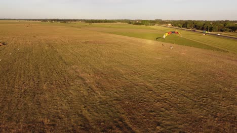 Luftaufnahme-Eines-Mehrfarbigen-Paratrike-Gleitschirms,-Der-Bei-Sonnenuntergang-über-Eine-Grüne-Wiese-Fliegt