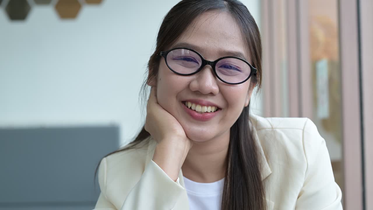 Close-up of happy young Asian businesswoman wearing eyeglasses looking through window then turning to camera and smiling happily