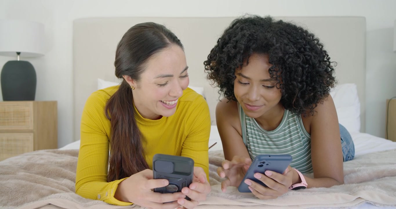 Diverse female friends lying on bed, scrolling phones, comparing posts and showing pink smartwatch