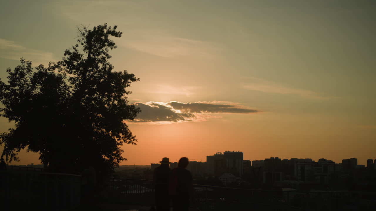 어두워지면서 철의 레일링 근처의 길을 따라 는 사람들의 실루, 배경에 도시 풍경, 큰 나무가 그림자를 던지고, 따뜻한 황금색 빛이 도시 스카이 라인을 비추고