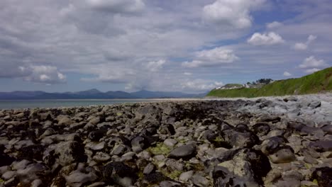 A-slow-Right-to-Left-smooth-Drone-track-across-a-rocky-beach-to-darker-rocks-with-distant-mountain-in-the-distance-in-Wales-in-the-UK---10-second-version