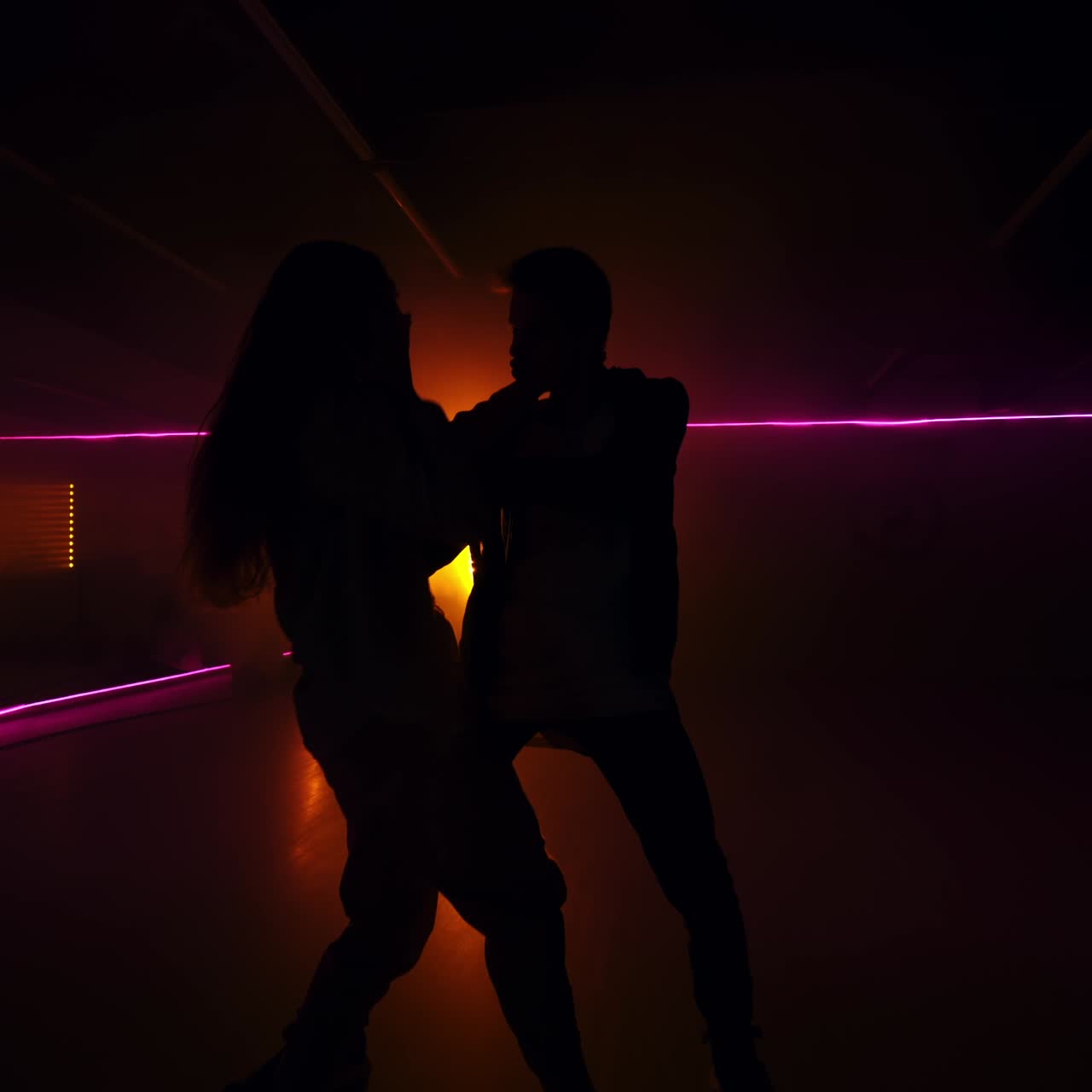 Silhouettes of a couple moving at the music. Dancers perform contemporary dance style in dark studio. Yellow light lamp at backdrop