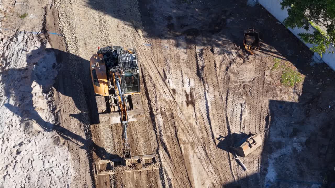 Excavator moves soil at construction site, aerial perspective, bright daylight, steady overhead camera movement