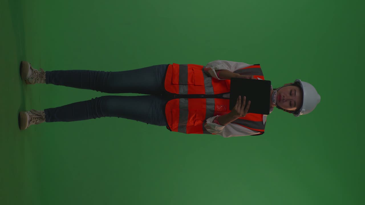 Full Body Of Asian Female Engineer With Safety Helmet Working On A Tablet While Standing In The Green Screen Background Studio, During Sunset Or Sunrise Time