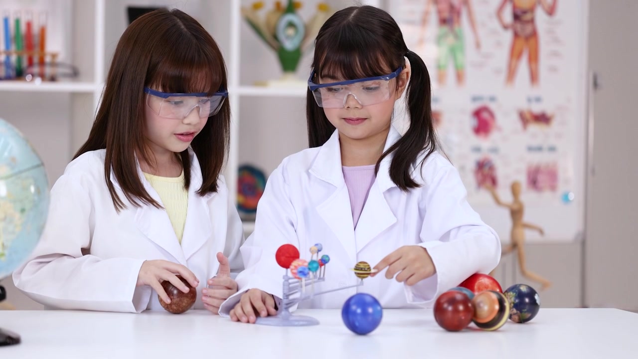 Two girls wearing safety goggles examine a planetary model in a bright science classroom setting.