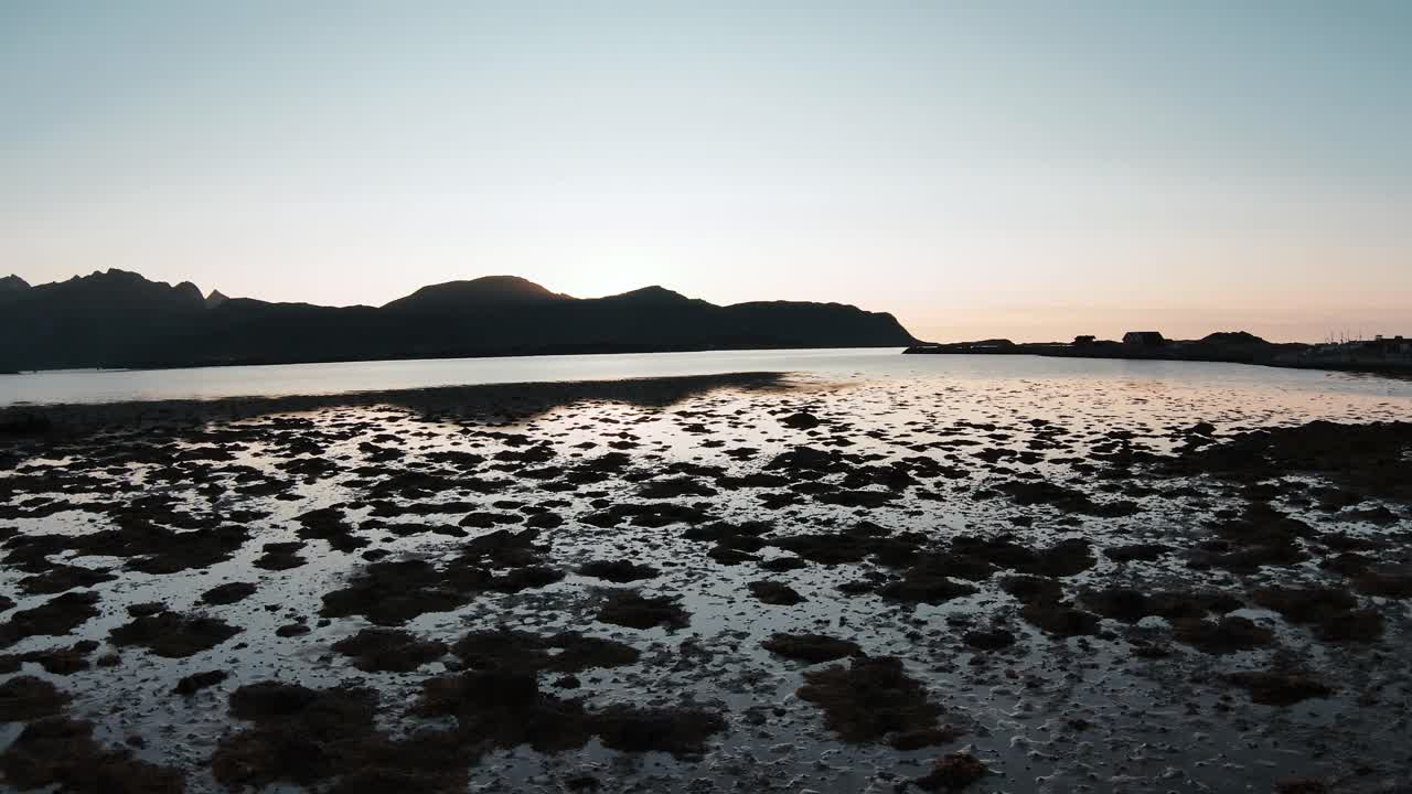 얕은 물, 배경에 있는 산, 그 뒤에 있는 태양을 슬로모션으로 바라보며 땅 가까이 해안을 날고 있다