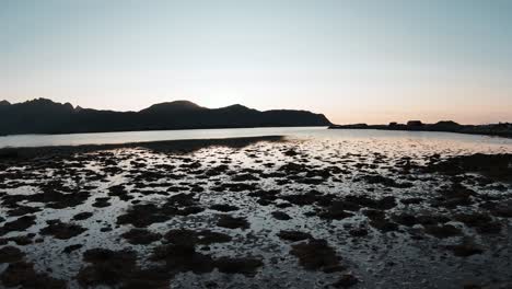 flying at the shore close to the ground looking at the shallow water, the mountains in the background and the sun setting behind them in slowmotion