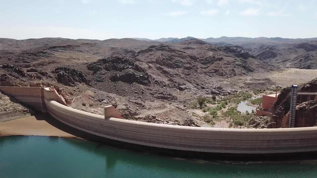 Vista aerea di una diga curva che trattiene un bacino idrico turchese nella regione di Ouarzazate, circondata da aspre montagne desertiche. Ideale per infrastrutture, gestione delle risorse idriche e immagini ambientali