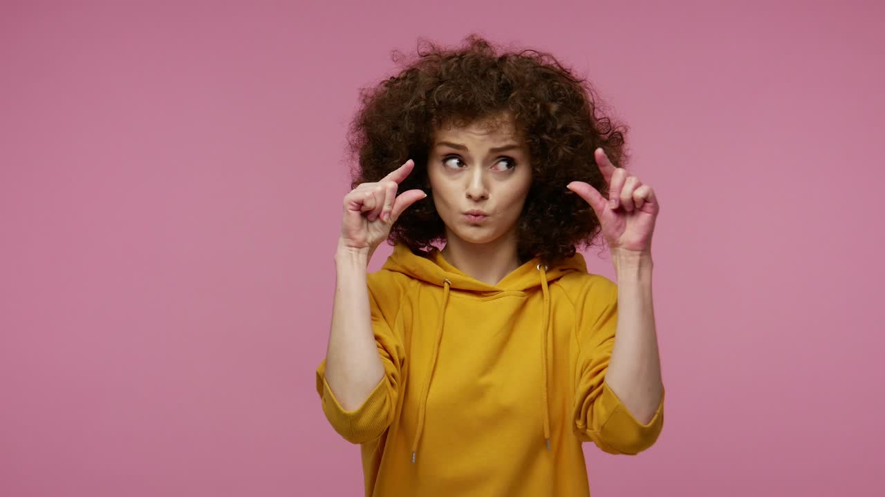 ¡demasiado pequeño! peinado afro de chica con capucha mostrando un pequeño signo, mirando con una mueca implorante y decepcionado por el tamaño