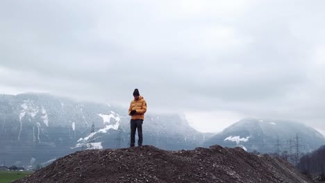 Drone-Aéreo-Orbitando-A-Un-Hombre-Con-Chaqueta-De-Invierno-Parado-En-Un-Montón-De-Grava-En-La-Naturaleza-Y-Disfrutando-De-La-Vista-Durante-El-Día-Nublado