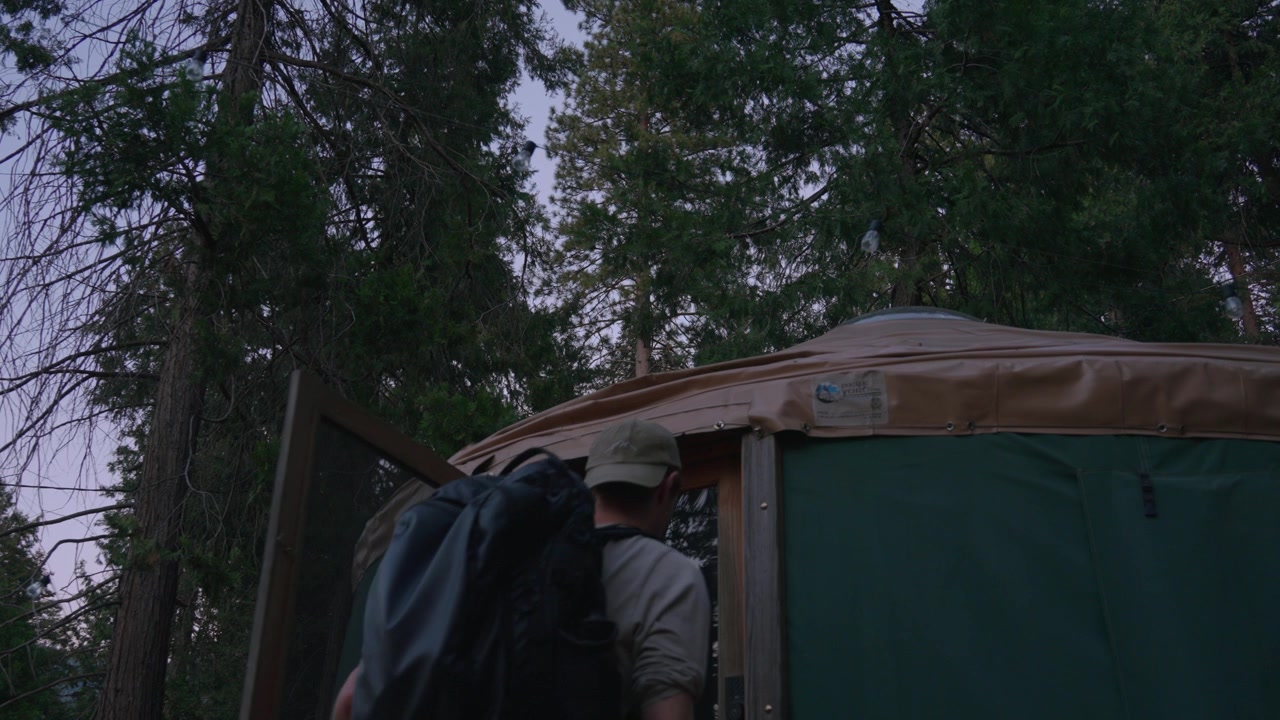 A hiker with a backpack walks past a green canvas yurt tent in a pine forest.