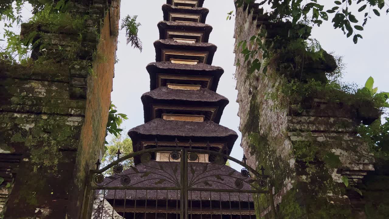 Traditionel balinesisk hinduhelligdom set gennem en udsmykket metalport, indrammet af ældgamle murstenstempelvægge og forvitrede stensøjler, omgivet af frodigt grønt på Bali, Indonesien.