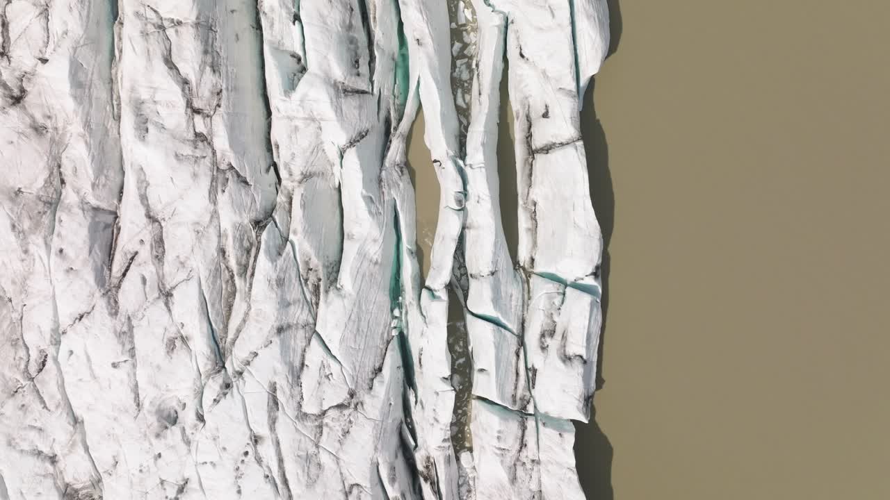 Top-down aerial view of Fjallsjökull Glacier’s fractured edge meeting the muddy waters of Fjallsarlon Lagoon, Iceland, showcasing sharp crevasses.