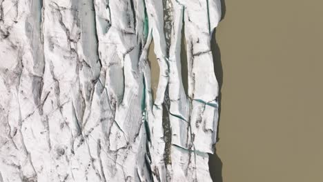 top-down aerial view of fjallsjökull glacier’s fractured edge meeting the muddy waters of fjallsarlon lagoon, iceland, showcasing sharp crevasses.