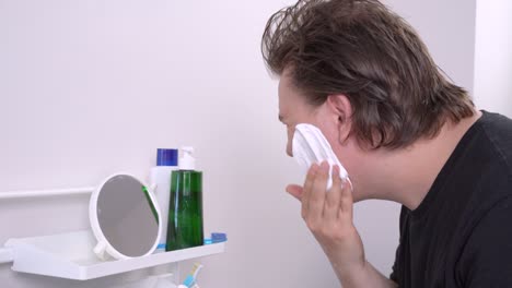 Daily morning hygiene procedures. Man washes with water, examines face in small mirror, applies shaving foam to cheeks and chin. Cosmetic cleansers, moisturizer and toothbrush on shelf in bathroom