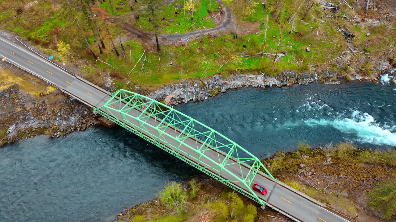 Quick river of sea-blue color flowing under the little metal bridge. Red car goes by the highway over the river. Aerial perspective.