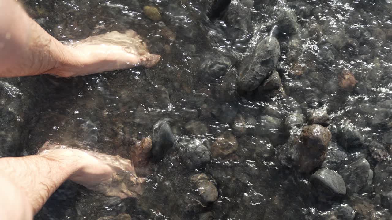 Man Standing on Tenerife Beach with Pebbles While Water Flows Around his Legs and Feet with a POV