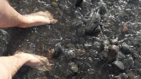 Man Standing on Tenerife Beach with Pebbles While Water Flows Around his Legs and Feet with a POV