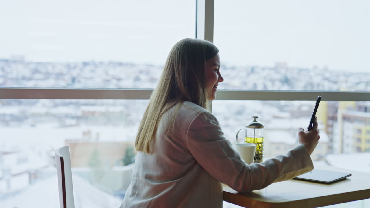 Happy smiling woman holding smartphone in front of herself and having a video call. Side view of a lady having tea break and relax.